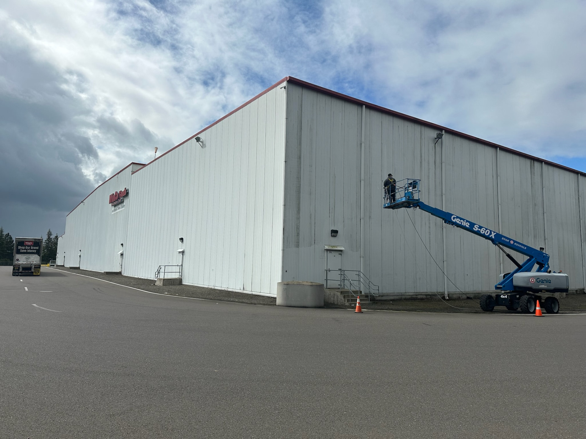 Technician washing a large industrial warehouse exterior with a boom lift